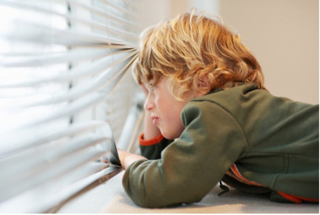 A boy peering out the binds covering a window.