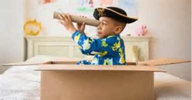 A boy dressed up in a pirate hat sitting in a carboard box looking through a paper towel tube.