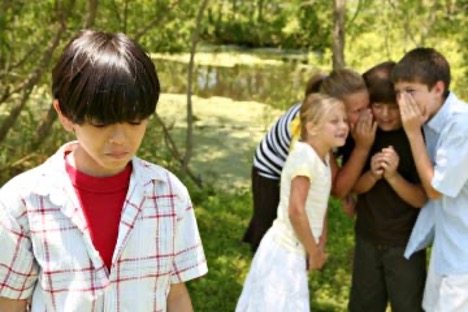 A group of children are whispering  together away from a child who has his head down and looking sad.