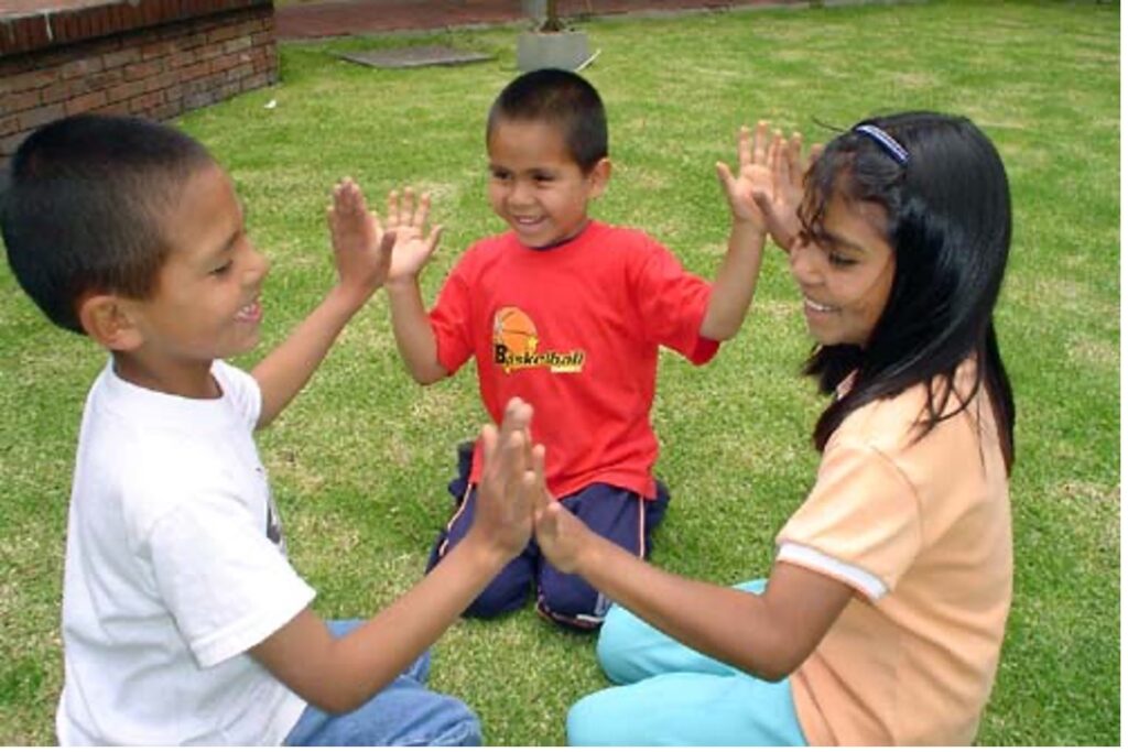 Three children sitting in a circle playing a patty cake game together with smiles on faces.