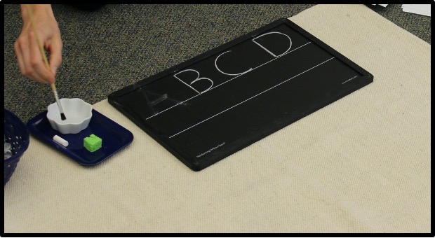 child using a wet paintbrush to trace letters on a chalkboard